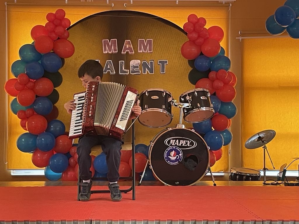 Jeden z uczestników konkursu szkolnego "Mam talent" grający na akordeonie, uczeń siedzi na krześle na scenie, w tle perkusja oraz napis "Mam talent".